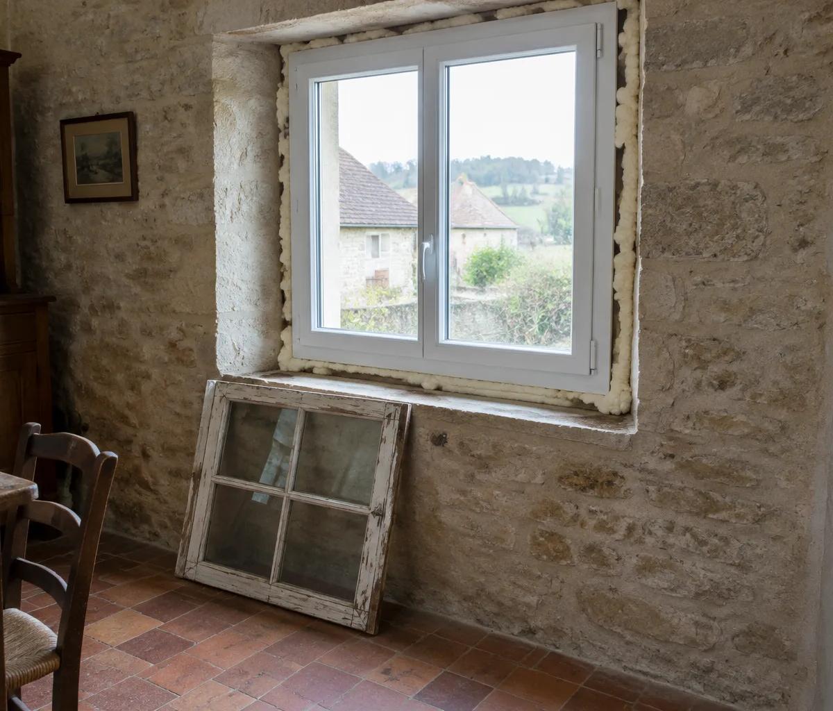 Fenêtre double vitrage neuve posée dans un mur en pierre, l'ancienne fenêtre en bois appuyée en dessous